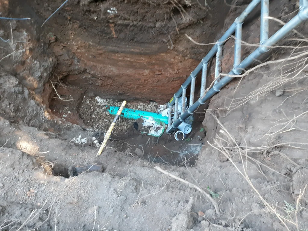 Excavated dirt trench with ladder and blue pipe.